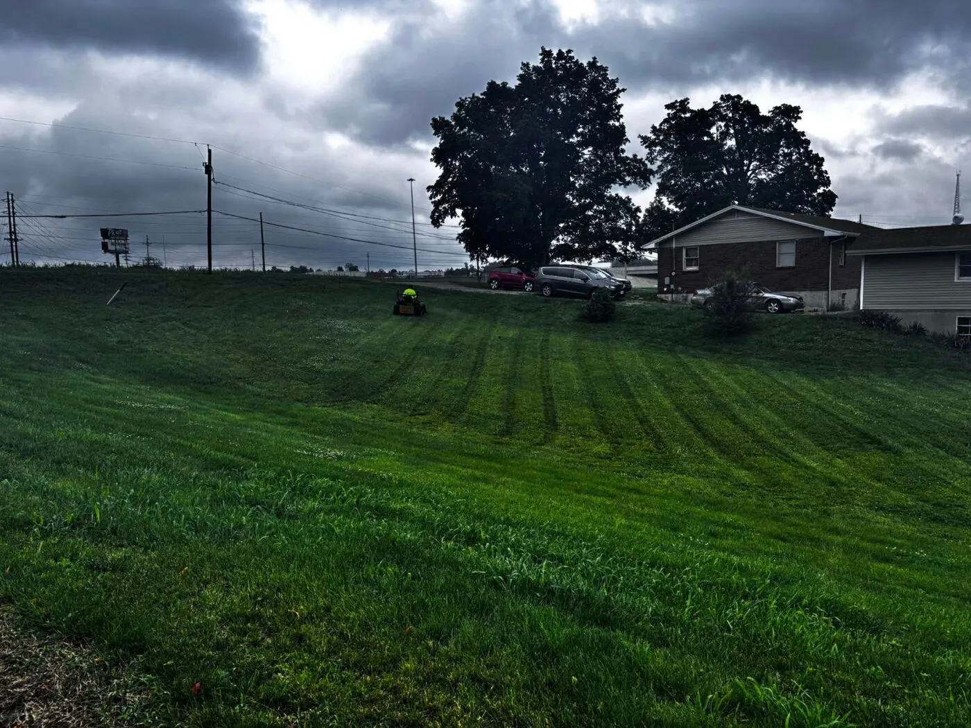 Freshly mowed hillside lawn on a cloudy day with a zero-turn mower and houses in the background.